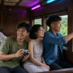 Four young Thai friends laughing by the open windows of a vintage train travelling through green countryside.