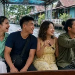 Young Thai friends riding a fast wooden commuter boat along a Bangkok canal, one lifting the side splash tarp