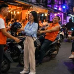 Young Thai passengers and riders in orange vests at a motorbike taxi stand at the entrance of a Bangkok soi